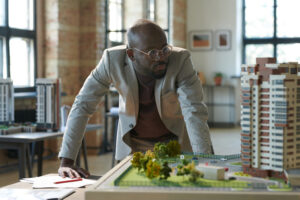 Serious architect in eyeglasses looking at construction model of new residential area during his work at office